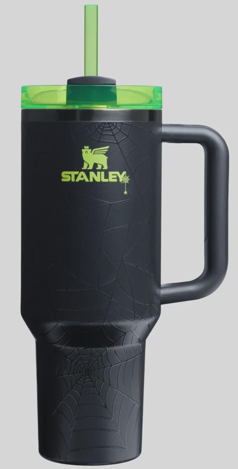 May include: Black Stanley tumbler with a green lid and straw. The tumbler features a spiderweb design and the Stanley logo in green. The handle is black. A Halloween-themed drinkware item.