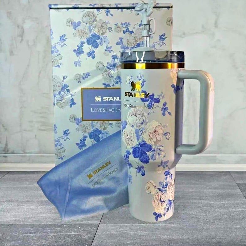 Op de afbeelding: Stanley-beker met bloemenpatroon, handvat, rietje en deksel, gepresenteerd met een bijpassende geschenkdoos en -tas. De beker heeft een lichte achtergrond met blauwe en witte bloemmotieven, gouden accenten en het Stanley-logo.