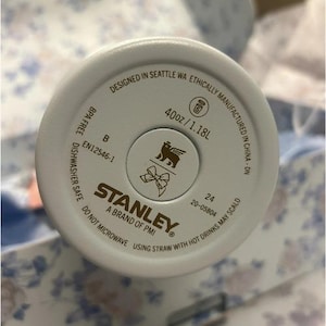 Op de afbeelding: Een close-up van de bodem van een witte Stanley-beker. De basis bevat de tekst "Designed in Seattle WA", "40oz/1.18L" en "BPA Free". Het Stanley-logo staat in het midden.