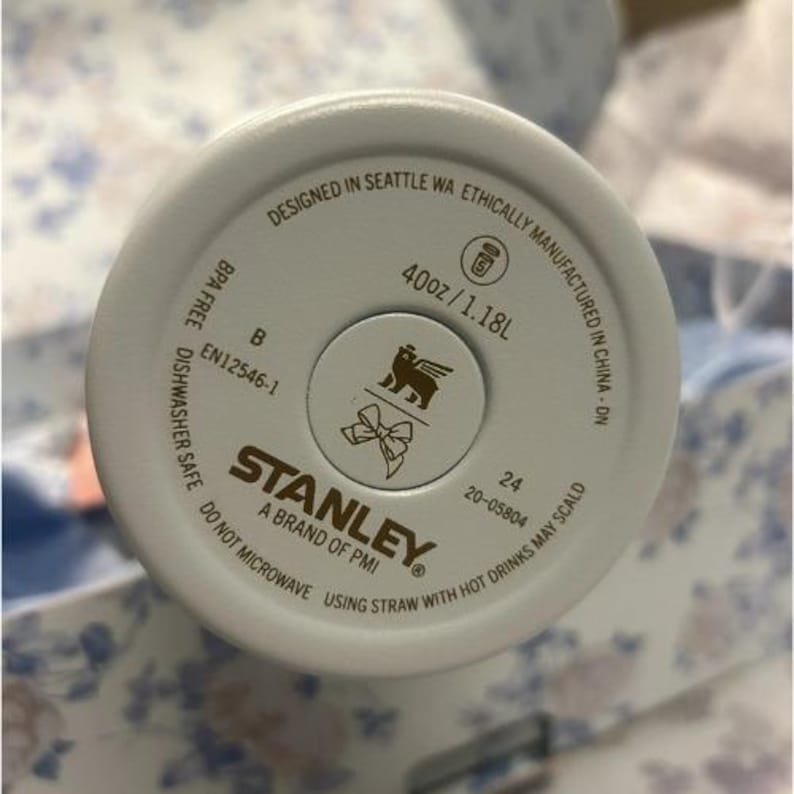 Op de afbeelding: Een close-up van de bodem van een witte Stanley-beker. De basis bevat de tekst "Designed in Seattle WA", "40oz/1.18L" en "BPA Free". Het Stanley-logo staat in het midden.