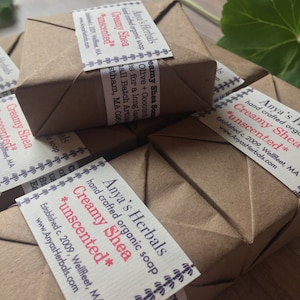 Puede incluir: Una pila de barras de jabón orgánico hecho a mano envueltas en papel marrón, etiquetadas con "Anya's Herbals Creamy Shea *unscented*" con la dirección del sitio web de la empresa y la fecha de establecimiento.