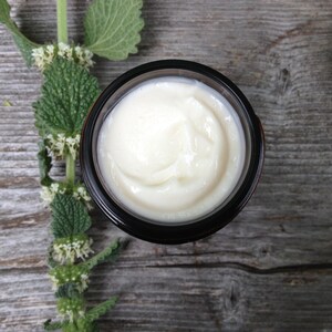May include: A jar of cream with a light, off-white color, set against a rustic wooden surface. The cream's texture is smooth and appears freshly applied. A sprig of green leaves and small white flowers is placed to the left of the jar.