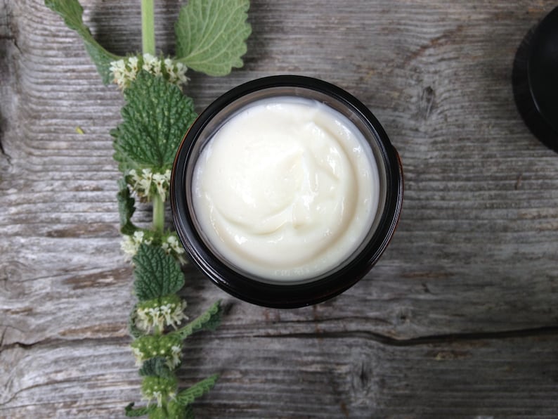 May include: A jar of cream with a light, off-white color, set against a rustic wooden surface. The cream's texture is smooth and appears freshly applied. A sprig of green leaves and small white flowers is placed to the left of the jar.