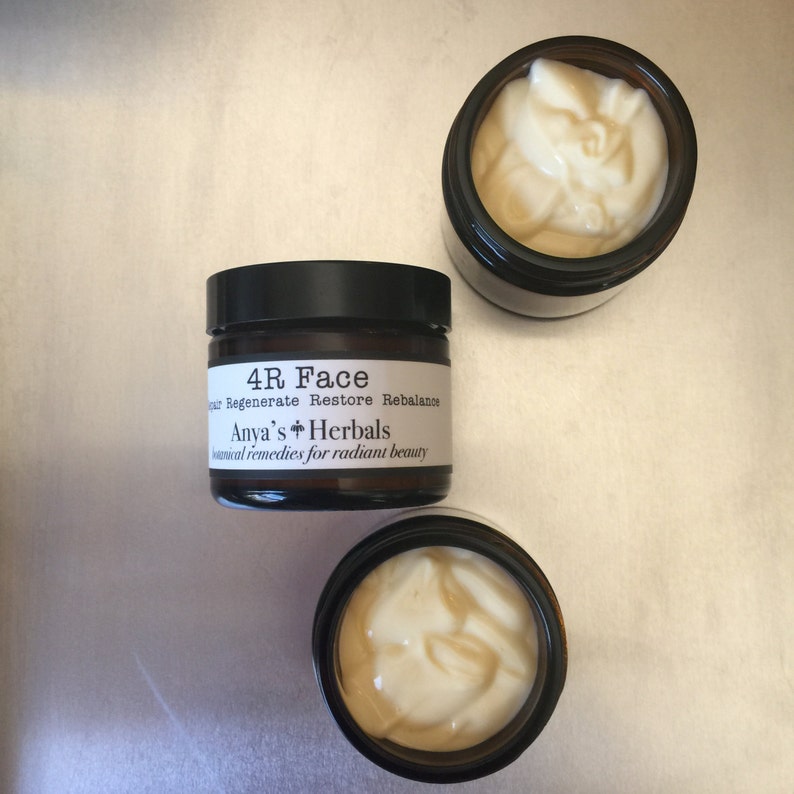 May include: Three small, dark brown glass jars with black lids, filled with a creamy, off-white face cream. The center jar has a label that reads "4R Face" and "Anya's Herbals, botanical remedies for radiant beauty."