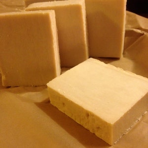 May include: Five rectangular bars of handmade soap, all the same size and shape, are arranged in a row on a brown paper surface. The soap is a light beige color and has a smooth, slightly textured surface.