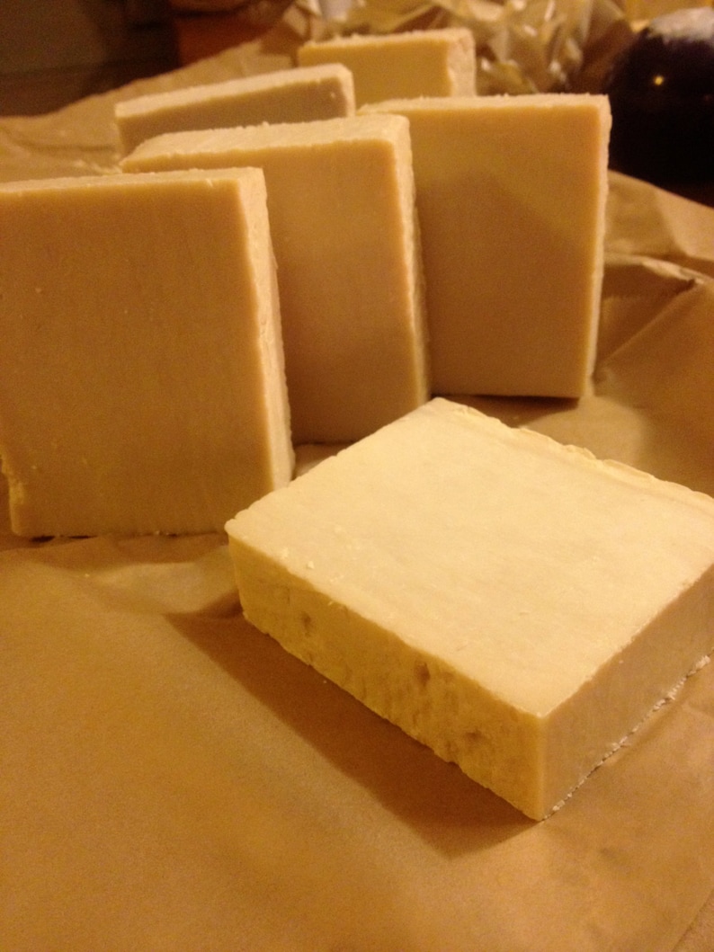 May include: Five rectangular bars of handmade soap, all the same size and shape, are arranged in a row on a brown paper surface. The soap is a light beige color and has a smooth, slightly textured surface.