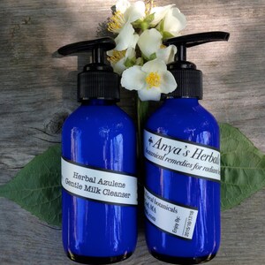 May include: Two blue bottles of Herbal Azulene Gentle Milk Cleanser by Anya's Herbal, botanical remedies for radiant skin. The bottles have black pump tops and white labels with black text.