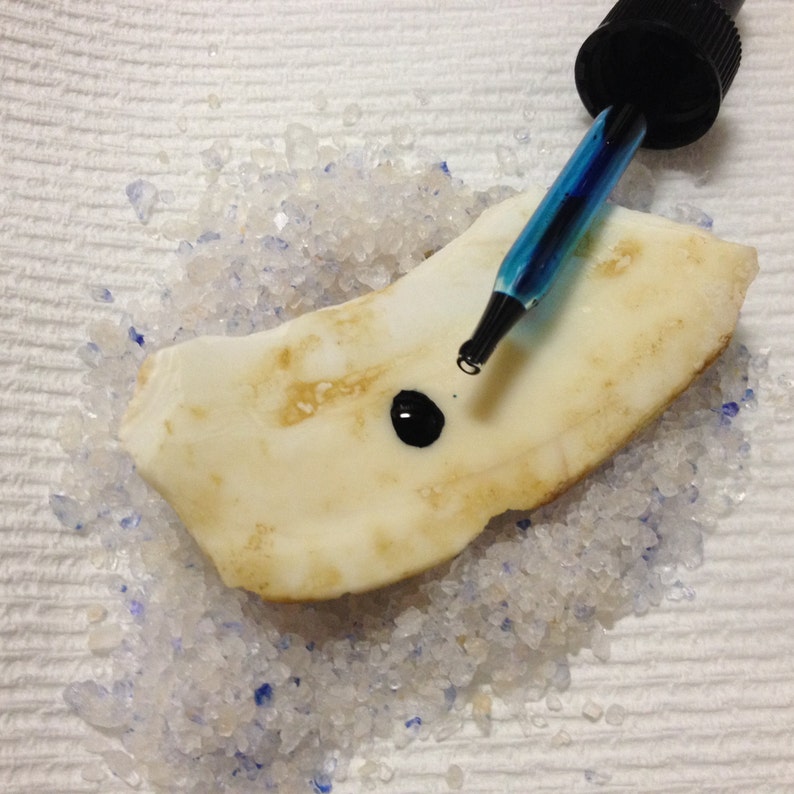 May include: A close-up of a white, irregularly shaped object with a small black drop of liquid on it. A blue dropper with a black cap is positioned above the object. The object is surrounded by small, white crystals.