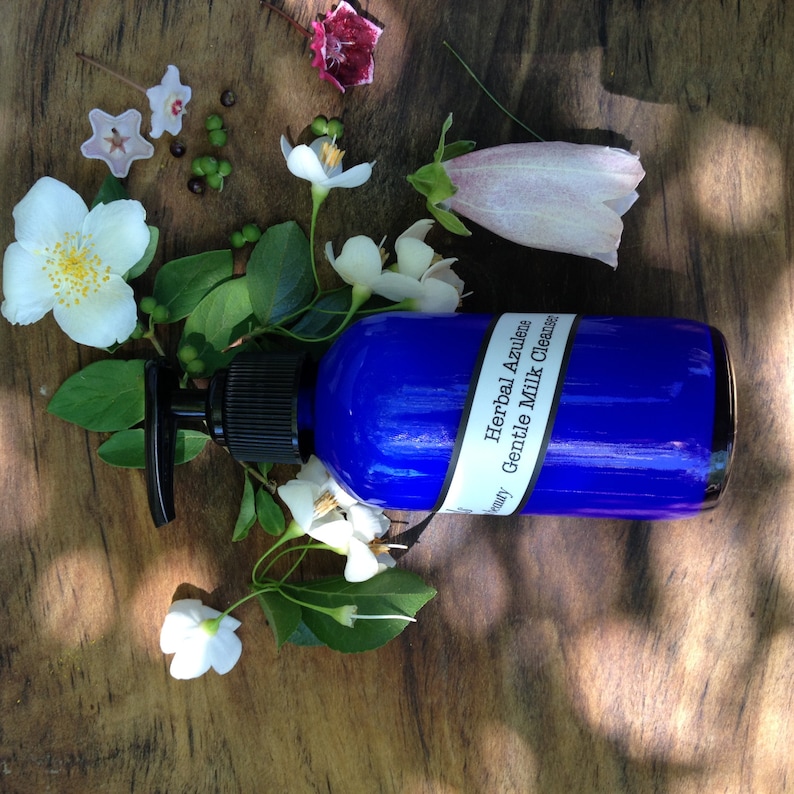May include: A blue bottle with a pump dispenser containing Herbal Azulene Gentle Milk Cleanser. The bottle is surrounded by white flowers and green leaves on a wooden surface.