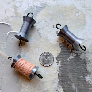 Puede incluir: Tres carretes de hilo grises y plateados con ganchos negros, uno con hilo color melocotón, se muestran sobre un fondo texturizado y abstracto. Un cuarto de dólar estadounidense está junto a los carretes. Los carretes son probablemente para coser o hacer manualidades.