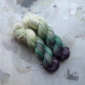 Può includere: Due gomitoli di filo, uno bianco e uno verde con accenti viola e bianchi. Il filo è disposto in un nodo sciolto.