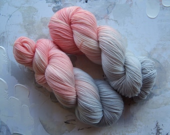 Puede incluir: Dos madejas de hilo en degradado de color, que transicionan de un melocotón suave a un gris claro. El hilo está dispuesto sobre una superficie texturizada y pintada con pinceladas blancas y grises, creando una exhibición visualmente atractiva para proyectos de manualidades.