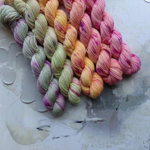 May include: A collection of colorful yarn skeins arranged diagonally. The yarn transitions from light green with purple accents to yellow, orange, and finally, vibrant pink with purple speckles. The background is a textured, abstract design.
