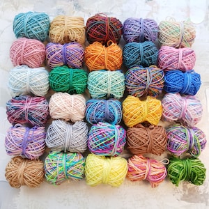 May include: An overhead shot of a collection of colorful yarn balls. The yarn balls are in various colors, including blue, pink, green, orange, and yellow. The yarn is tightly wound and appears to be soft and fluffy. The background is a light, textured surface.