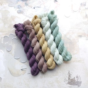 May include: Five skeins of yarn in shades of purple, brown, yellow, green, and light green. The yarn is arranged in a gradient from dark to light.