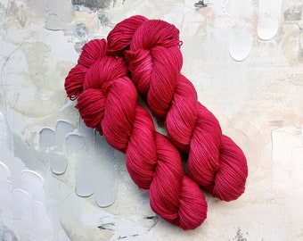 Puede incluir: Madeja de hilo fucsia vibrante, bien torcido y listo para tejer. El hilo se muestra sobre un fondo texturizado y abstracto con trazos de pintura blancos y grises. Ideal para proyectos de punto o ganchillo.