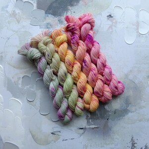 May include: A collection of colorful yarn skeins arranged in a diagonal pattern. The yarn features a gradient of colors, including pink, yellow, orange, and green. The background is a textured, abstract surface with white paint strokes.