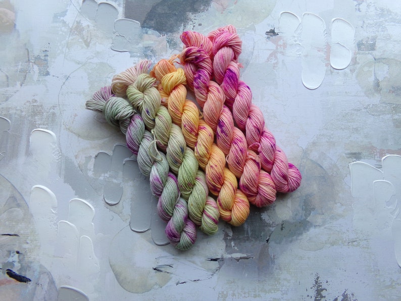 May include: A collection of colorful yarn skeins arranged in a diagonal pattern. The yarn features a gradient of colors, including pink, yellow, orange, and green. The background is a textured, abstract surface with white paint strokes.