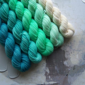 May include: A collection of yarn skeins in a gradient of colors, transitioning from deep teal to light green and cream. The yarn is arranged in neat rows, showcasing the color variations. The background has a textured, neutral surface.
