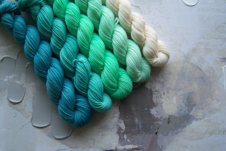 Puede incluir: Madejas de hilo en un degradado de colores, que van desde el verde azulado intenso hasta el verde claro y el crema. El hilo est&aacute; dispuesto en filas ordenadas, mostrando las variaciones de color. El fondo tiene una superficie texturizada y neutra.