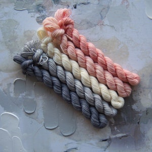 May include: A collection of six skeins of yarn in a gradient of colors, from dark gray to light pink. The yarn is arranged diagonally against a textured, off-white and gray background.