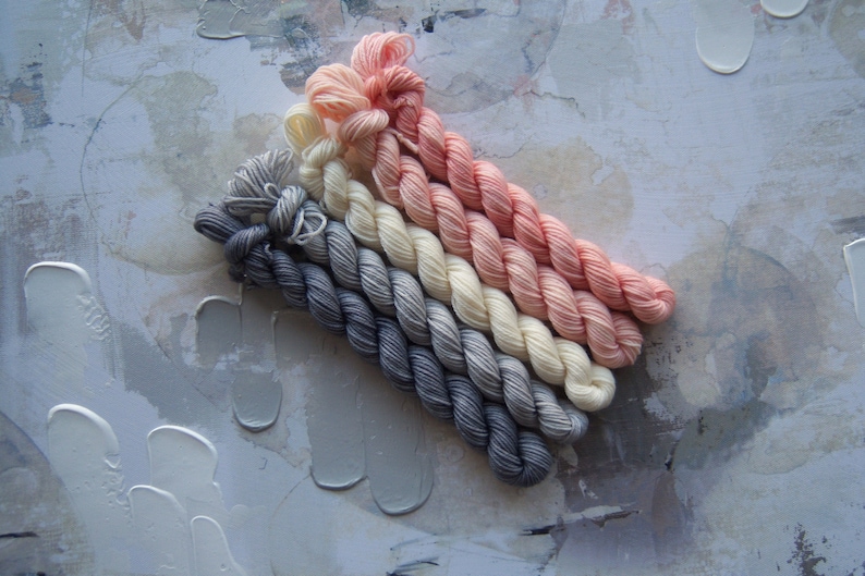 May include: A collection of six skeins of yarn in a gradient of colors, from dark gray to light pink. The yarn is arranged diagonally against a textured, off-white and gray background.