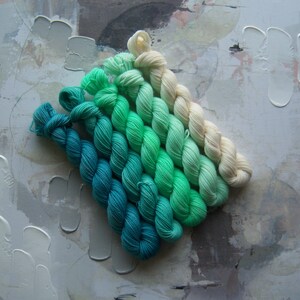 Puede incluir: Una colecci&oacute;n de madejas de hilo en un degradado de colores, que van desde el verde azulado intenso hasta el crema claro. El hilo est&aacute; dispuesto en diagonal sobre un fondo texturizado de tonos neutros, mostrando las vibrantes transiciones de color.