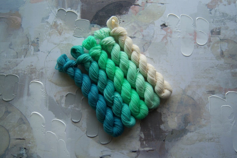 Puede incluir: Una colecci&oacute;n de madejas de hilo en un degradado de colores, que van desde el verde azulado intenso hasta el crema claro. El hilo est&aacute; dispuesto en diagonal sobre un fondo texturizado de tonos neutros, mostrando las vibrantes transiciones de color.