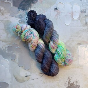 Pode incluir: Dois novelos de fio, um &eacute; de cor cinza escuro e o outro &eacute; um fio multicolorido com tons de verde, rosa e branco.