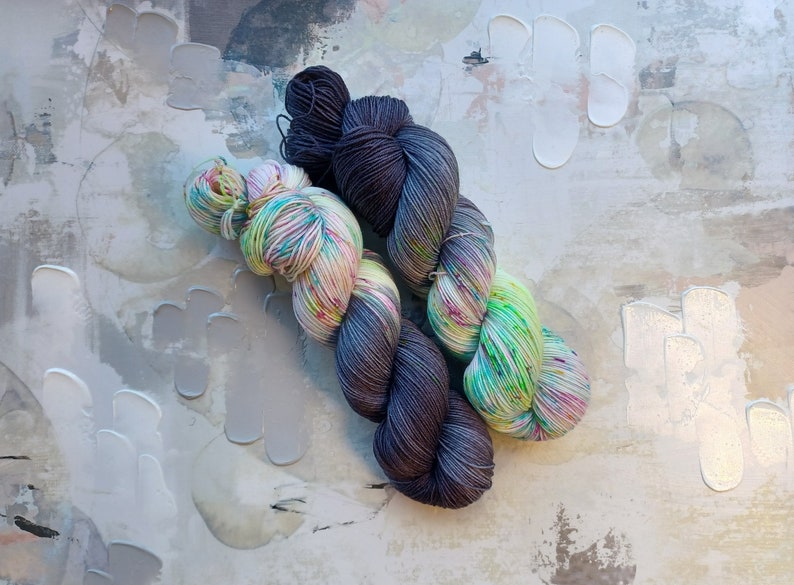 Pode incluir: Dois novelos de fio, um &eacute; de cor cinza escuro e o outro &eacute; um fio multicolorido com tons de verde, rosa e branco.