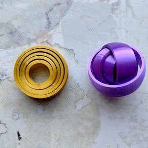 May include: Two 3D-printed objects. One is a yellow, concentric ring design. The other is a purple sphere with interwoven bands. The objects are on a textured, off-white surface.