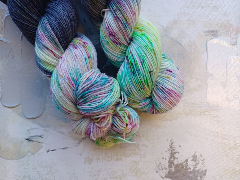 Pode incluir: Dois novelos de fio, um &eacute; um degrad&ecirc; azul escuro e cinza, o outro &eacute; uma base branca com respingos de rosa, verde e azul.