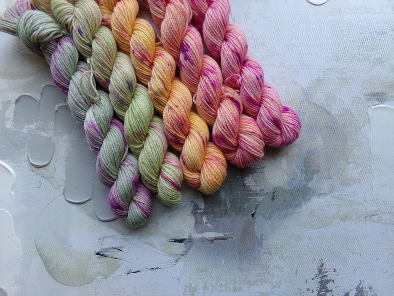 May include: A collection of colorful yarn skeins arranged diagonally. The yarn transitions from light green with purple accents to yellow, orange, and finally, vibrant pink with purple speckles. The background is a textured, abstract design.