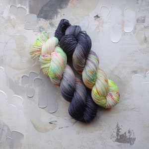 Pode incluir: Dois novelos de fio, um &eacute; cinza claro com um efeito degrad&ecirc; cinza escuro, o outro &eacute; um fio branco com respingos de cores brilhantes como rosa, verde e amarelo.