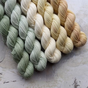May include: Close-up of several skeins of yarn in a gradient of colors, ranging from light green to cream and ending in a peach hue. The yarn is tightly wound and appears soft, ideal for knitting or crochet projects.