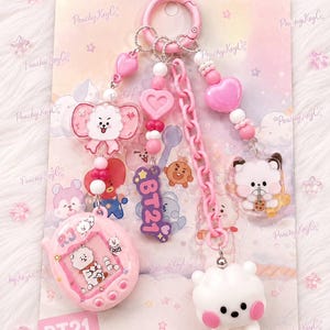 May include: A pink keychain featuring a white RJ character charm, a pink digital pet-style charm with RJ graphics, a purple BT21 logo charm, and a pink plastic chain. The keychain is adorned with various pink and white beads and heart-shaped accents.