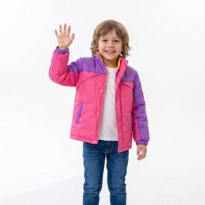May include: A colour-blocked puffer jacket featuring a bright pink main body and purple sleeves, shoulders, and collar. The jacket includes a yellow inner lining, a front zip closure with snap buttons, and two side pockets, worn over a white shirt and blue jeans.