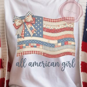 Puede incluir: Camiseta blanca con un diseño de bandera americana y un lazo. El diseño de la bandera utiliza patrones rojos, blancos y azules. El texto "all american girl" está impreso debajo. La persona lleva una rebeca con un estampado de estrellas y rayas.