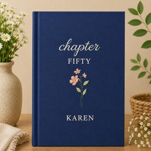 May include: A navy blue hardcover book with the words "chapter FIFTY KAREN" in white script. A floral embroidery design of pink flowers and green leaves is centered below the text. The book is on a wooden surface.