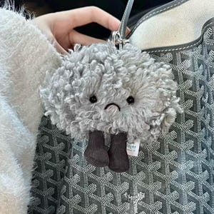 May include: A gray plush cloud-shaped keychain with a sad face and dark gray boots. The keychain has a silver clasp and a small tag that reads "LYCAT". The plush toy is held in front of a patterned tote bag.