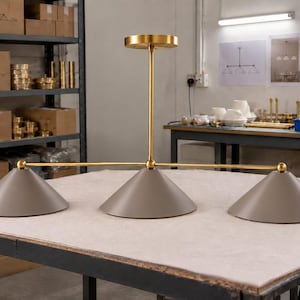 May include: A three-light pendant fixture featuring a horizontal brass rod connected to a central vertical stem. The fixture includes three matte taupe conical shades attached to the ends and center of the rod, finished with small brass spheres.