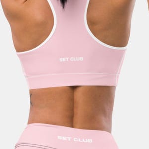 Puede incluir: Dos mujeres con conjuntos de entrenamiento a juego. Uno es rosa claro con ribete blanco, el otro es naranja con ribete blanco. Cada conjunto incluye un sujetador deportivo y leggings. El texto "SET CLUB" está impreso en los sujetadores deportivos y leggings.