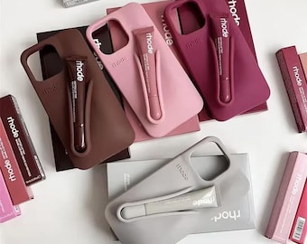 Könnte beinhalten: Vier Handyhüllen aus Silikon in Braun, Hellrosa, Dunkelrosa und Grau, jede mit einer geformten Halterung für eine passende Rhode Peptide Lip Tint Tube, präsentiert zusammen mit den entsprechenden Rhode-Produktverpackungen.
