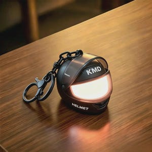 May include: A black motorcycle helmet keyring featuring a translucent visor, a black chain, and a circular ring. The helmet has the text KMD printed in white on the upper section and the word HELMET printed in white on the lower front section.