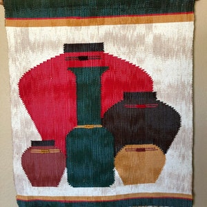 May include: Woven wall hanging featuring a design of stylized vases in shades of red, green, brown, and tan. The artwork has a white background with a red circular shape. The top and bottom edges have decorative stripes and fringe.