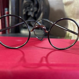 May include: Round eyeglasses with a dark brown frame and gold accents. The lenses are clear, and the bridge of the glasses is a darker brown. The glasses are resting on a red surface.