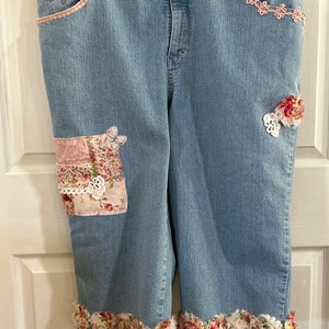 May include: Light blue denim capri pants with decorative floral patches and lace trim. The capris feature a pink lace trim on the pockets and a floral ruffle hem. The pants are hanging on a wooden hanger.