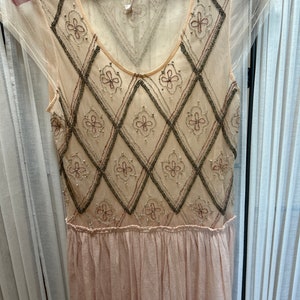 Op de afbeelding: Een mouwloze, cr&egrave;mekleurige jurk met een doorschijnende overlay. De bodice heeft een ruitpatroon met geborduurde bloemaccenten. Het onderste deel van de jurk is zachtroze, geplooid.