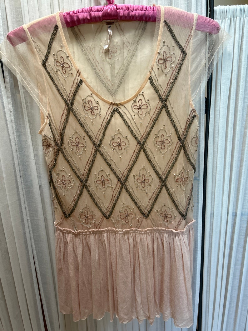 Op de afbeelding: Een mouwloze, cr&egrave;mekleurige jurk met een doorschijnende overlay. De bodice heeft een ruitpatroon met geborduurde bloemaccenten. Het onderste deel van de jurk is zachtroze, geplooid.