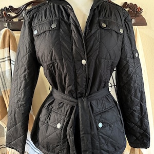 May include: Black quilted jacket with a belted waist. The jacket features a stand-up collar, silver-tone buttons, and multiple pockets. The diamond-patterned stitching adds a classic touch to this outerwear piece.